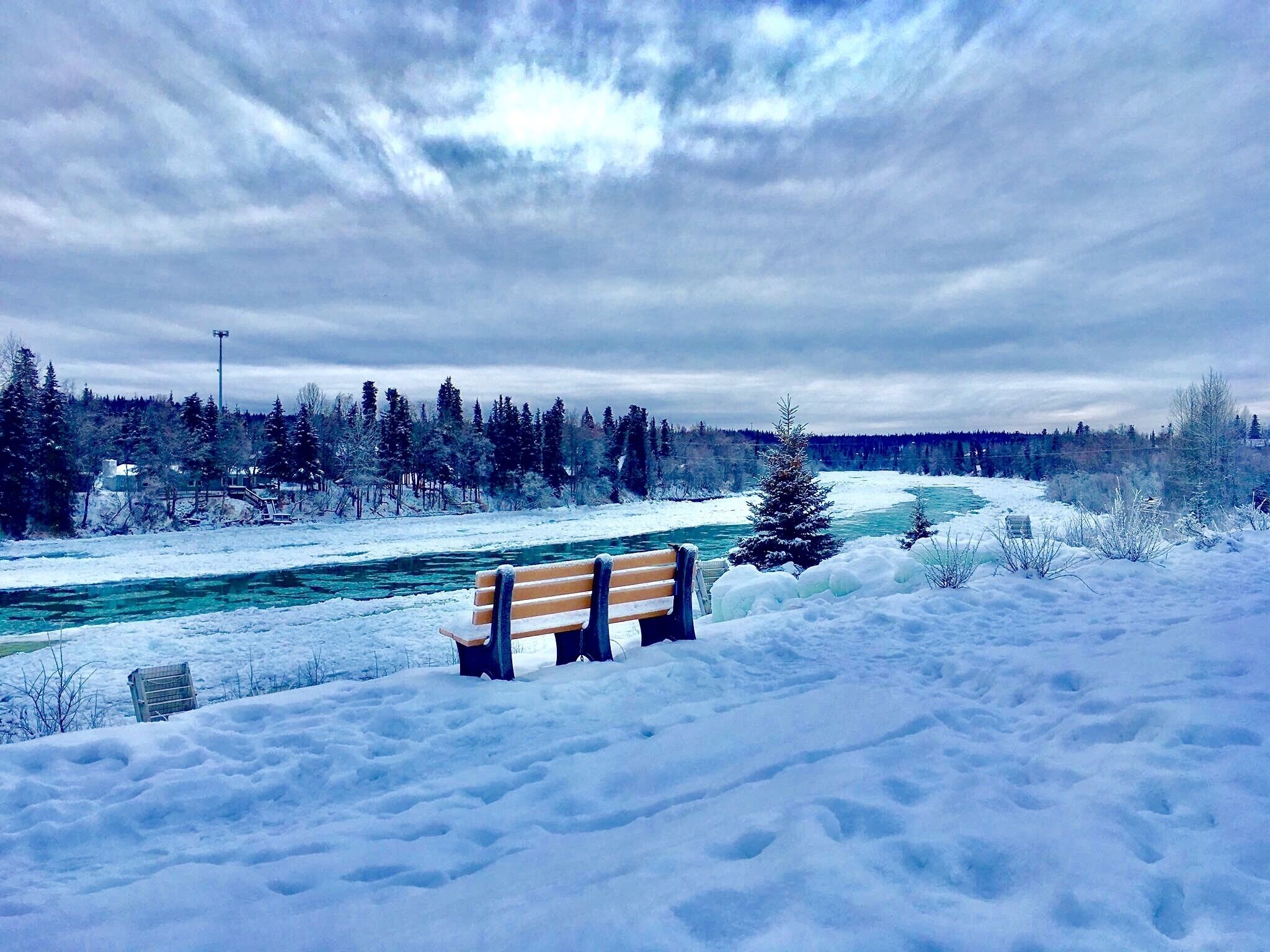 A snow-covered landscape with a bench, a river, and a forest.