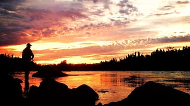La silhouette d’une personne se tenant sur des rochers au bord d’une rivière, au coucher du soleil.