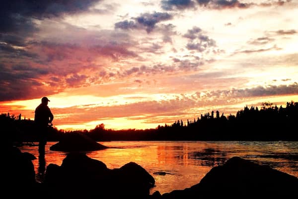 A silhouette of a person standing on rocks by a river at sunset.