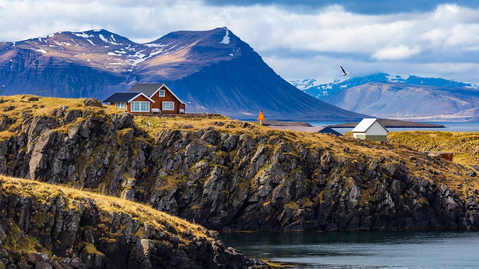 A coastal landscape with a house on a rocky hill, a mountain in the background, and a body of water in the foreground.