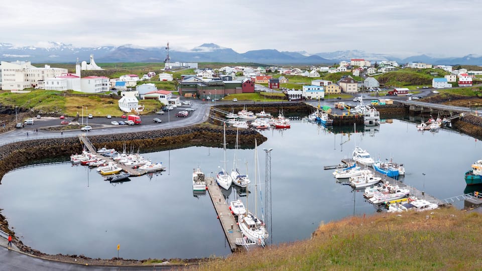A harbor with boats, a road, and buildings in a coastal town with mountains in the background.