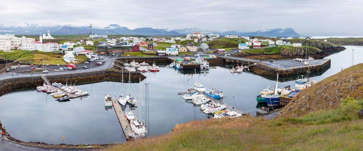 A harbor with boats, a road, and buildings in a coastal town with mountains in the background.