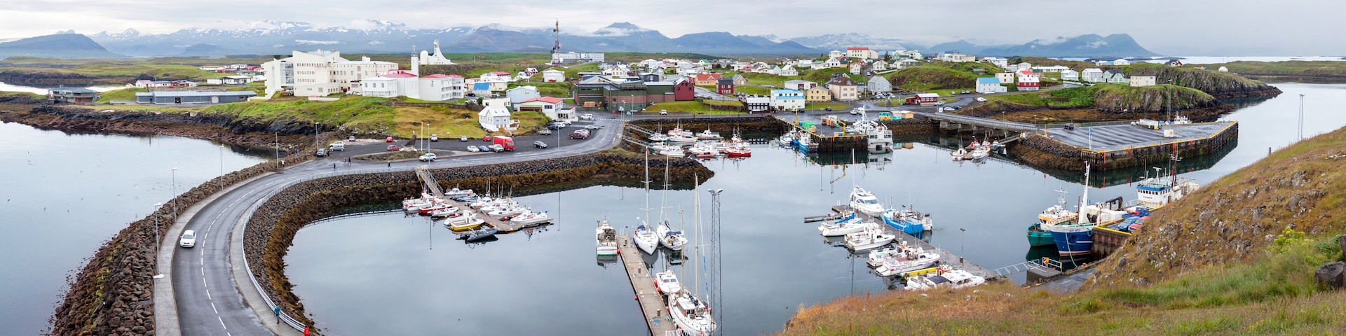 A harbor with boats, a road, and buildings in a coastal town with mountains in the background.