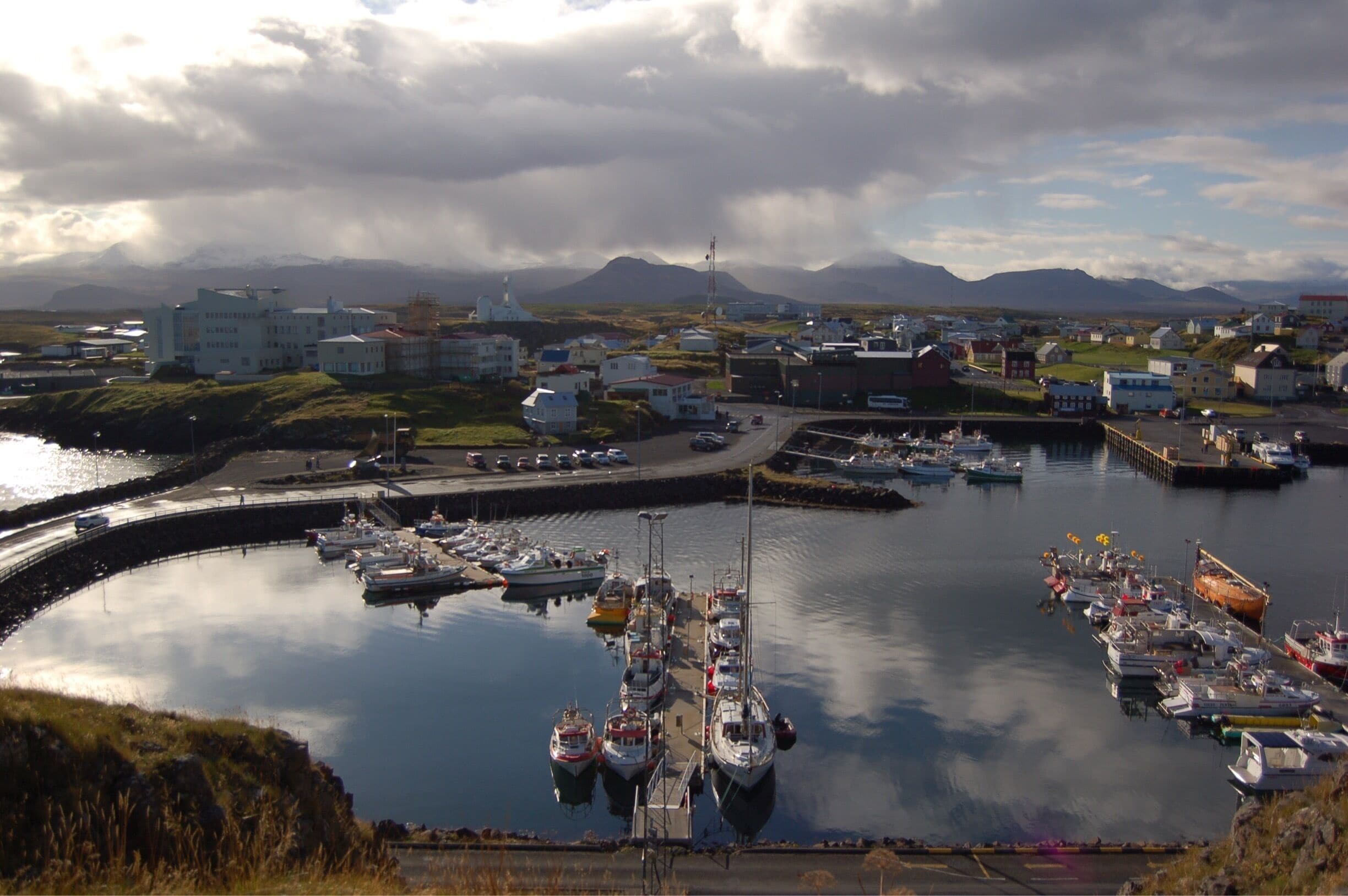 A harbor with boats docked, a marina, and buildings in the background with mountains.