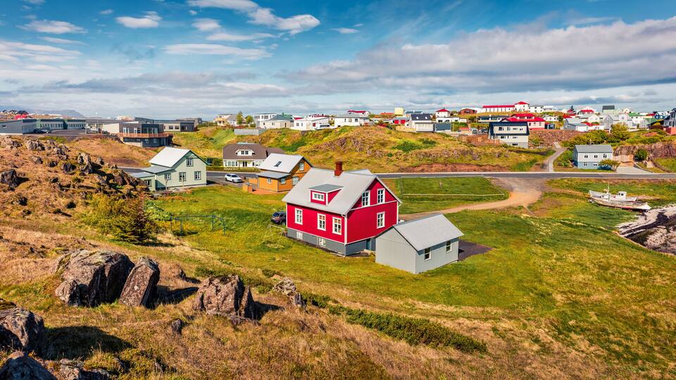 A coastal village with red and white houses, a grassy hill, and a rocky shoreline.