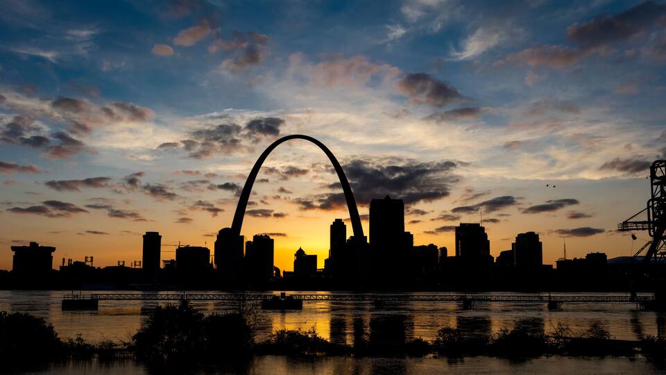 A city skyline at sunset with a prominent arch bridge silhouetted against the sky.
