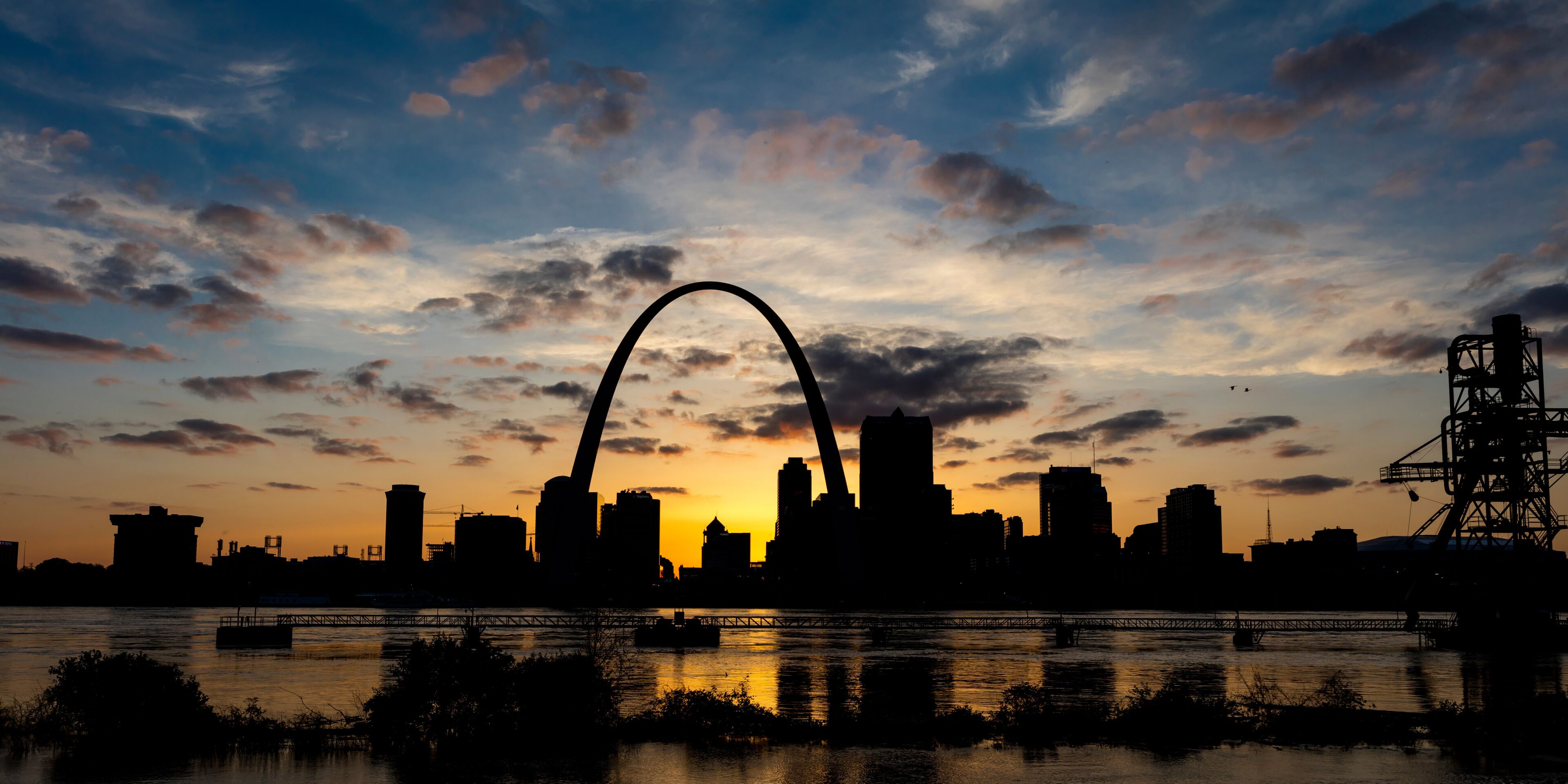 A city skyline at sunset with a prominent arch bridge silhouetted against the sky.