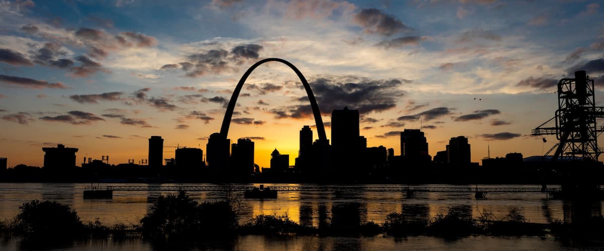 A city skyline at sunset with a prominent arch bridge silhouetted against the sky.