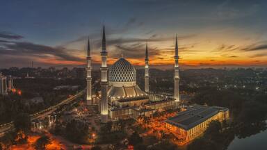 Uma mesquita com quatro minaretes e uma cúpula grande, rodeada por um horizonte urbano ao entardecer.