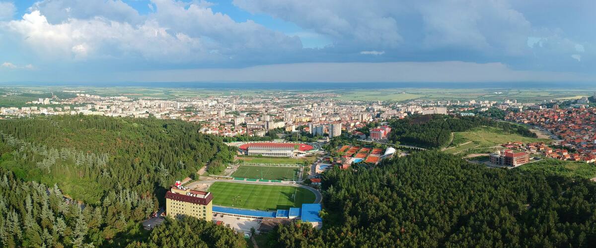 A cityscape with a stadium surrounded by dense forests and a distant view of a larger city.