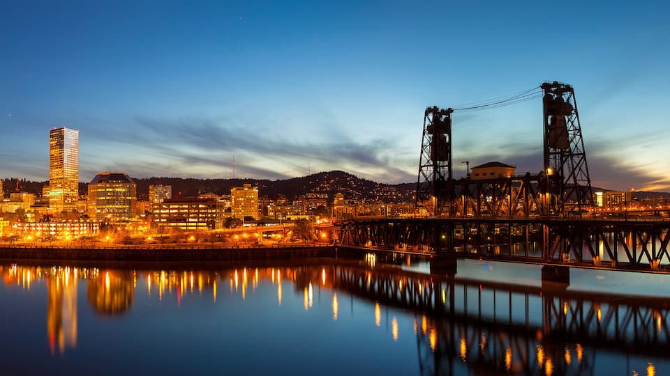 A city skyline at dusk with illuminated buildings reflecting on the water.