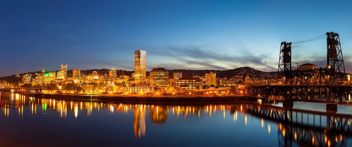 A city skyline at dusk with illuminated buildings reflecting on the water.