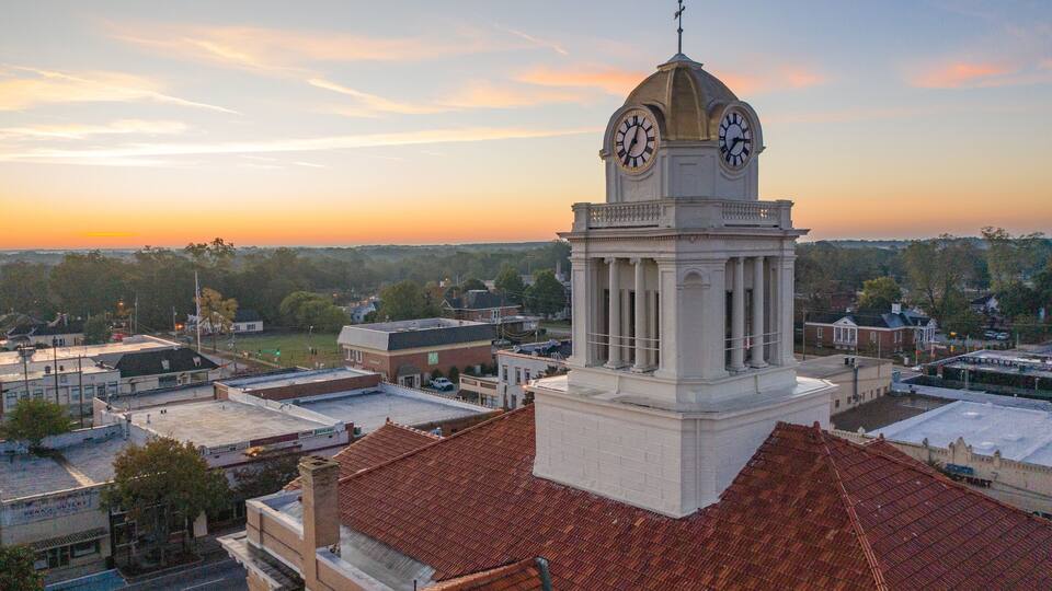 A clock tower with a dome and columns, surrounded by buildings and trees under a sunset sky.