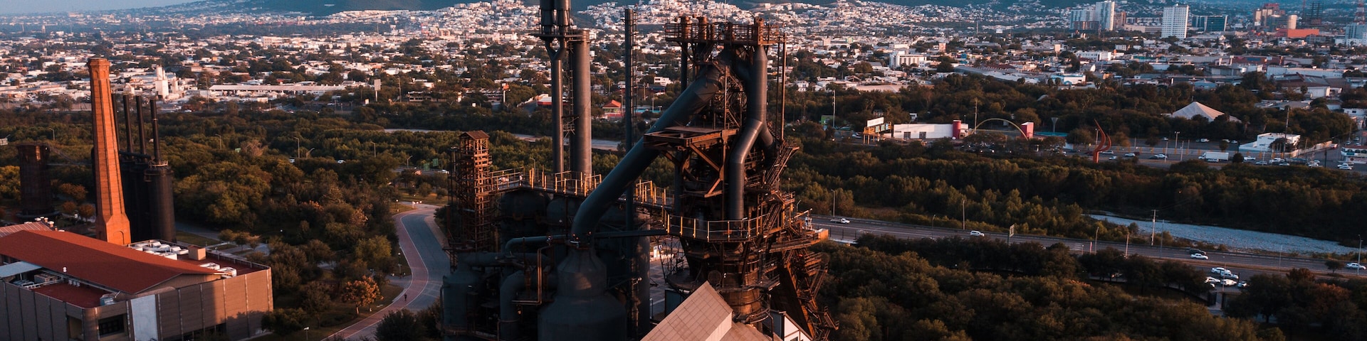 Un gran complejo industrial con múltiples chimeneas altas, rodeado por un paisaje urbano y una montaña al fondo.