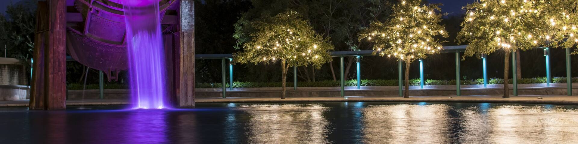 A nighttime scene with a water feature, illuminated trees, and a mountain in the background.
