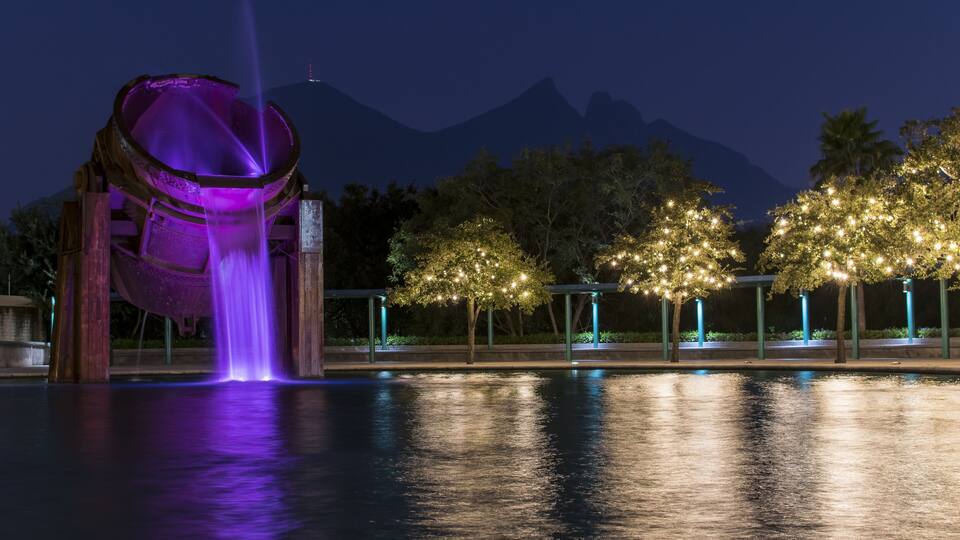 A nighttime scene with a water feature, illuminated trees, and a mountain in the background.