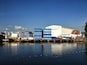 Un port situé au bord de l’eau, avec des conteneurs empilés et un grand bâtiment blanc.