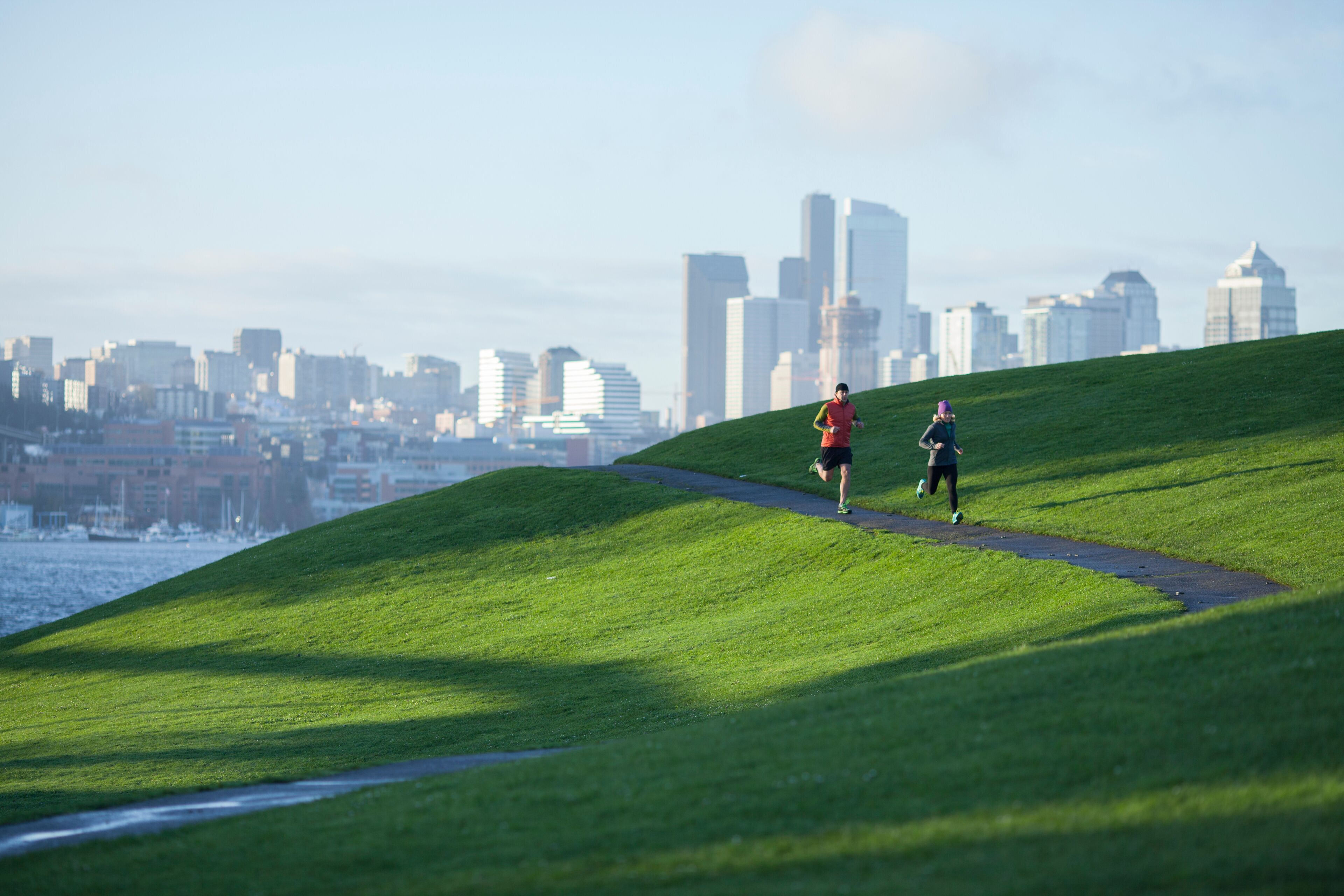 An adult man and woman running the paved path through Gasworks Park with the Seattle cityscape in the background. Washington State.