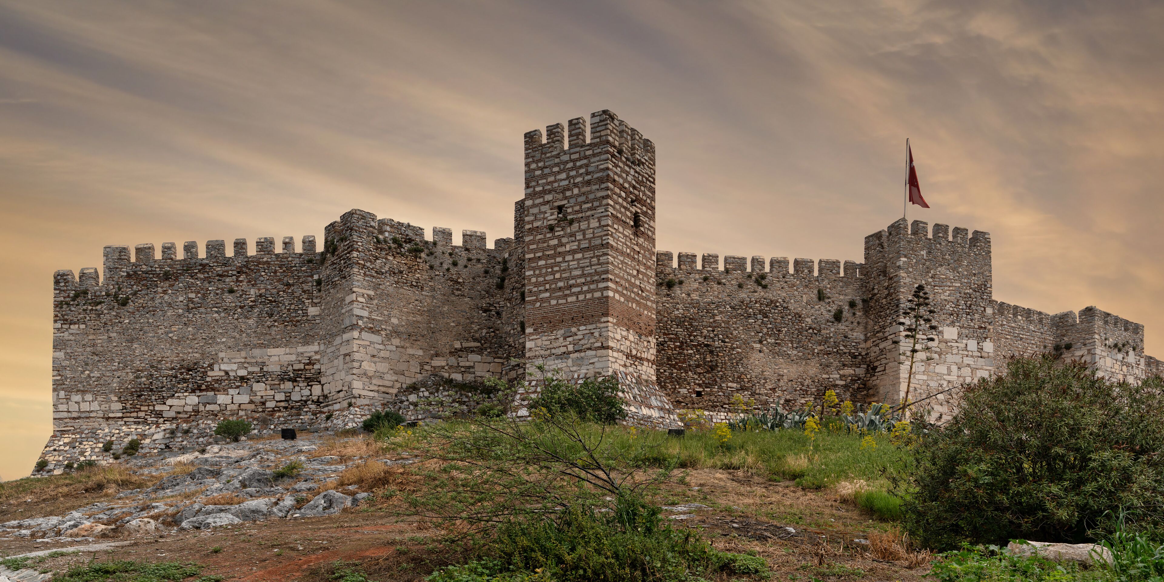 Ayasuluk Castle, Selcuk, Turkey. Ancient stone fortress walls, battlements, and a tower with the Turkish flag, seen from a rocky, vegetated hill against a cloudy sky