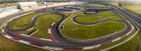 Concord, United States - 28 July 2025: Aerial view of a race track's winding asphalt curves cutting through vibrant green fields, contrasted by the stark white and red safety barriers.